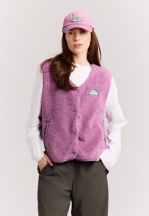Vrouw die een roze fleecevest en bijpassende pet draagt, witte lange mouwen shirt en donkere broek, staand met handen in de zakken.