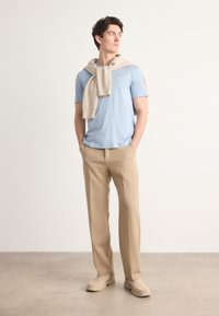 T-shirt azzurro chiaro a maniche corte, maglione beige a maglia appoggiato sulle spalle, pantaloni beige e scarpe slip-on beige chiaro su sfondo neutro.
