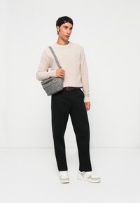 Man wearing beige sweater, black pants, white sneakers, and gray crossbody bag, standing against a white background.