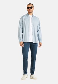 Hellblaues Hemd über einem weißen T-Shirt, kombiniert mit navyblauen Hosen und beigen Sneakern. Eine Sonnenbrille rundet das Outfit ab.