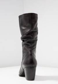 Bota de cuero negra hasta la rodilla con un diseño arrugado, que presenta un tacón apilado y una textura suave. Costuras visibles a lo largo del tubo.