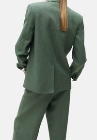 Traje verde a medida visto desde la parte de atrás, con una sola abertura, mangas largas con botones y una textura de tela suave y ligera.