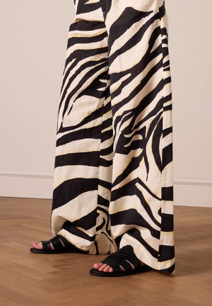 Pantalon ample en tissu à imprimé zèbre noir et blanc, avec des rayures audacieuses et une texture légère, associé à des sandales à lanières noires.