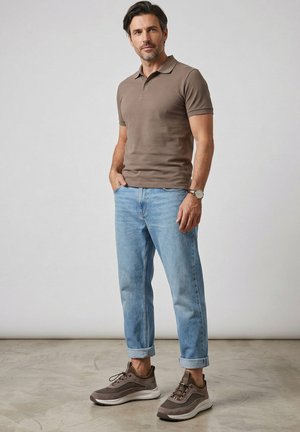 Muž stojící s rukama v kapsách, oblečený v hnědém polotričku, světle modrých ohrnutých džínách, hnědých teniskách a s hodinkami na zápěstí na šedém pozadí.