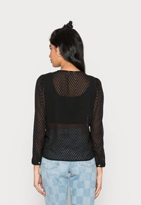 Blusa preta de mangas longas com um padrão em chevron texturizado, tecido semi-transparente e decote redondo. Combinada com calças xadrez azul claro.
