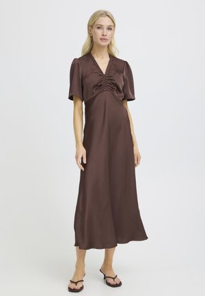 Blondynka ubrana w brązową, marszczoną sukienkę midi z krótkim rękawem oraz czarne sandały na obcasie, na jednolitym tle.