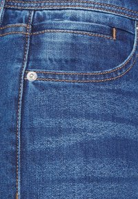 Mörkblå denimtyg med subtila texturerade mönster, en bakficka med en silverknapp och kontrasterande orange sömmar.