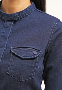 Tumma sininen farkkupaita, jossa on pystytakaulus, rintatasku napilla korostettuna, sileä pinta ja näkyvät ompelusaumat.