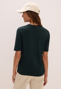 Dunkelgrünes kurzärmeliges T-Shirt aus weichem Stoff mit rundem Ausschnitt. Eine cremfarbene Baseballkappe ergänzt das Outfit.