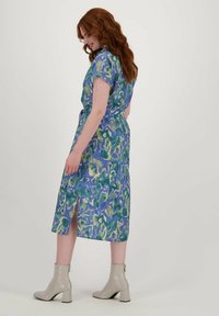 Korte mouw, knie-lange jurk in een abstract patroon in blauw en groen. Heeft een splitrok en een tailleband, gecombineerd met beige enkellaarsjes.