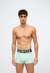 Muskuļots vīrietis ar īsiem cirtainiem matiem, ģērbies gaiši zilos Calvin Klein bokserīšos ar melnu jostu, stāv pret vienkāršu fona.
