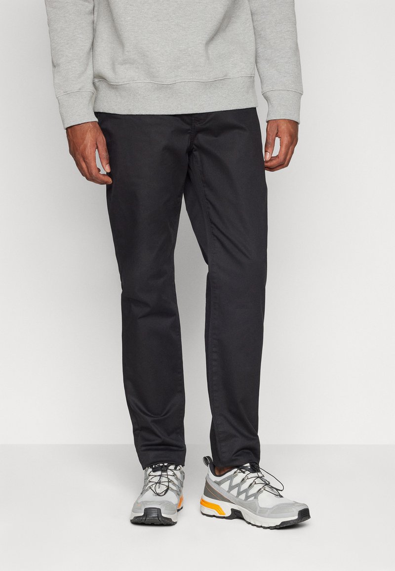 Pantaloni neri a taglio dritto, realizzati in un tessuto liscio, abbinati a scarpe sportive grigie e arancioni con superficie testurizzata e lacci.