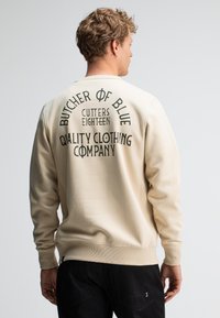Beige sweatshirt met lange mouwen, met zwarte tekst op de achterkant: "BUTCHER OF BLUE CUTTERS EIGHTEEN QUALITY CLOTHING COMPANY."