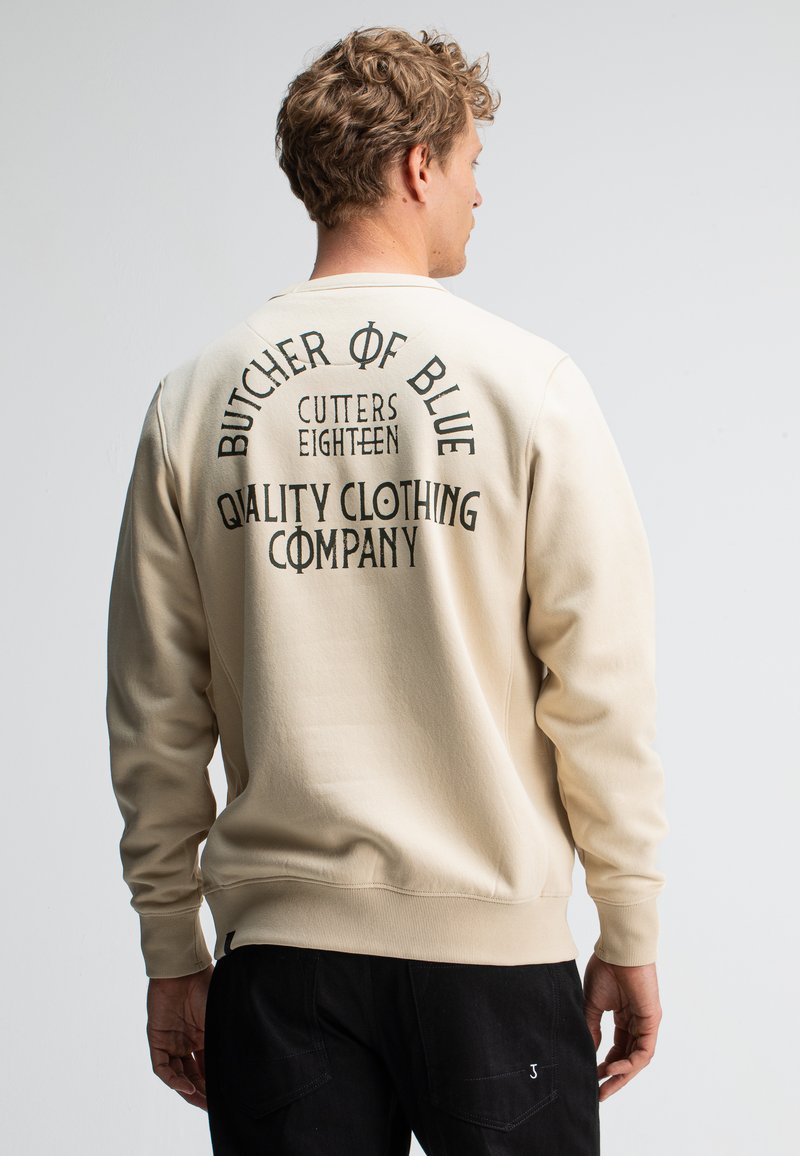 Beige sweatshirt met lange mouwen, met zwarte tekst op de achterkant: "BUTCHER OF BLUE CUTTERS EIGHTEEN QUALITY CLOTHING COMPANY."