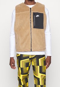 Tan fleeceväst med zipframsidan och ficka i svart med Nike-logotypen. Matchad med mönstrade gula och gröna byxor.