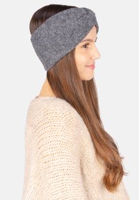 Un bandeau en feutre gris présente un design tressé, offrant chaleur et couverture. Un pull en laine beige tricoté est visible en arrière-plan.