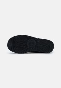 Semelle de chaussure noire avec motifs de bandes rayonnantes et en zigzag ainsi qu'un logo embossé au centre, photographiée sur un fond clair uni.