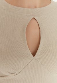 Haut beige côtelé avec un col rond et une ouverture en forme de goutte d'eau au centre devant. Texture douce avec une coupe ajustée et des détails froncés.