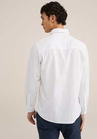 Witte langarmshirt met een button-down kraag, geplaatst op een lichte achtergrond; heeft een gebogen zoom en knoopdetails op de manchetten.