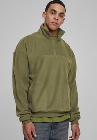 Pullover in pile verde oliva con collo a mezza zip e polsini a coste. Texture morbida, con una cucitura orizzontale sul petto.