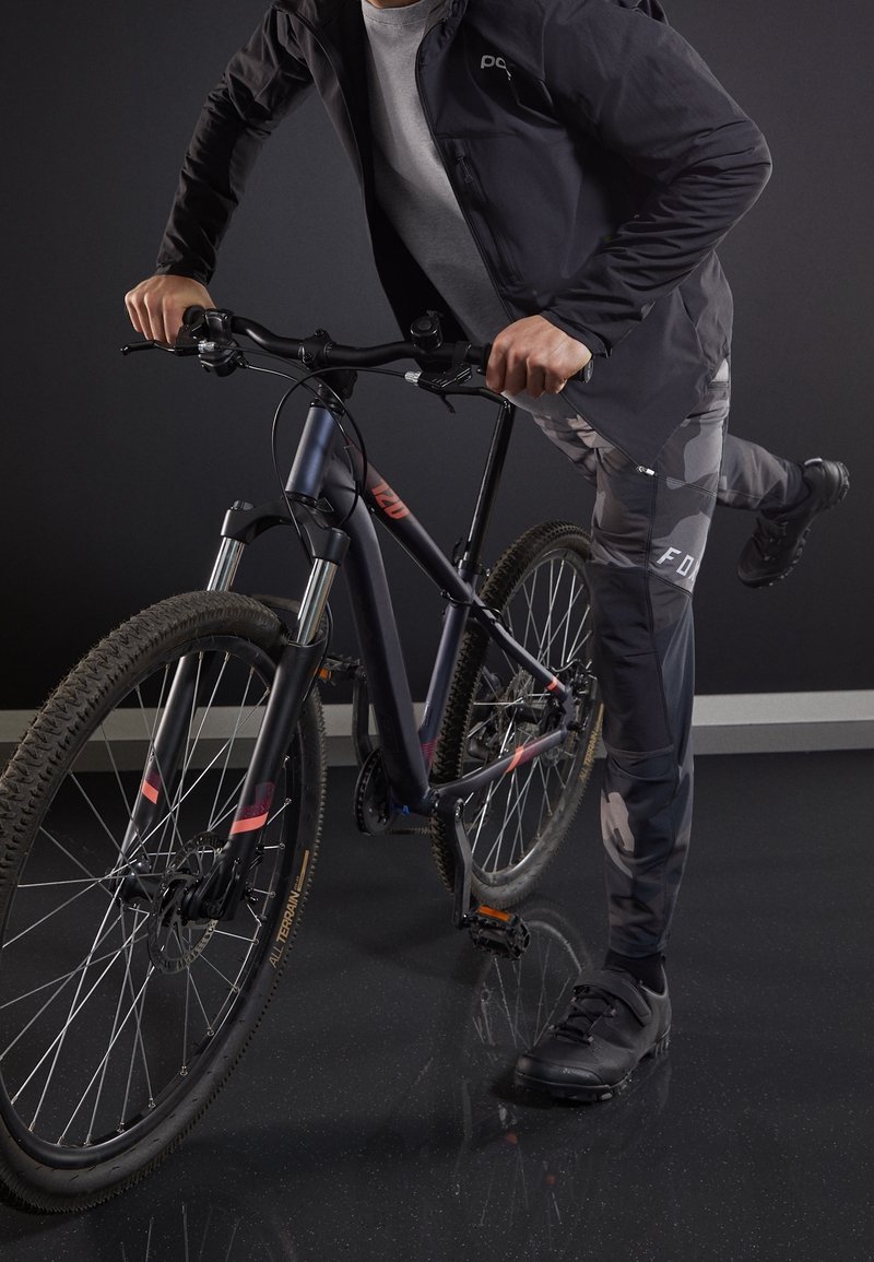 Mountain bike con telaio nero, forcelle di sospensione argento e pneumatici texturizzati; il ciclista indossa una giacca nera, pantaloni mimetici e scarpe nere.