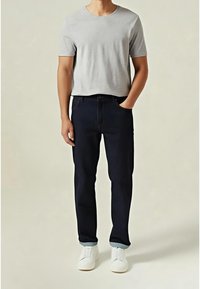 Mann trägt hellgraues T-Shirt mit kurzen Ärmeln, dunkelblaue Jeans mit Aufschlag am Saum, weiße Slip-on Schuhe, eine Hand in der Tasche, steht vor einfarbigem Hintergrund.