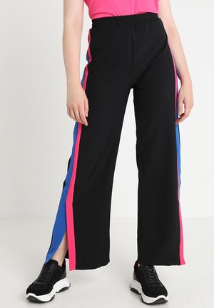 Persona che indossa pantaloni neri a gamba larga con strisce laterali rosa e blu e sneakers nere, in piedi su sfondo bianco.
