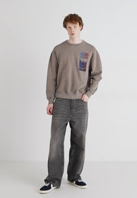 Felpa grigia con un piccolo patch grafico colorato sul lato sinistro del petto, abbinata a jeans grigi larghi e sneakers blu navy su uno sfondo semplice.