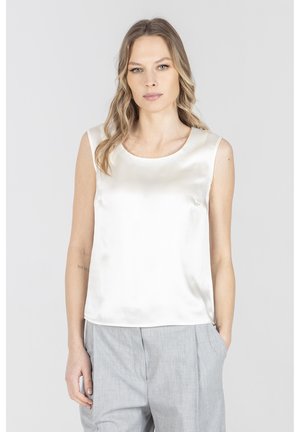 Kobieta o falowanych blond włosach, ubrana w bezrękawnik z białego satynowego topu i jasnoszare spodnie, stoi z jedną ręką w kieszeni na jednolitym tle.