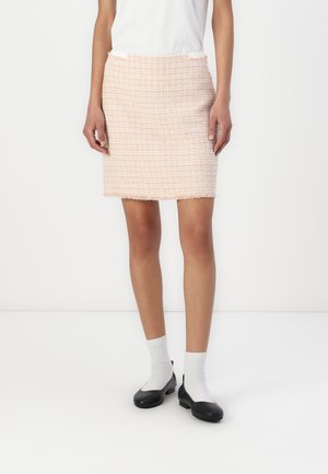 Femme portant une mini-jupe texturée rose clair, un haut blanc, des chaussettes blanches et des ballerines noires, debout sur un fond blanc.