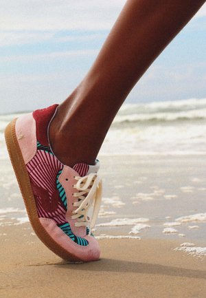 Pied posant sur du sable humide près de l'écume de mer, chaussé d'une basket colorée avec des motifs roses, rouges et bleus et des lacets blancs sous un ciel partiellement nuageux.