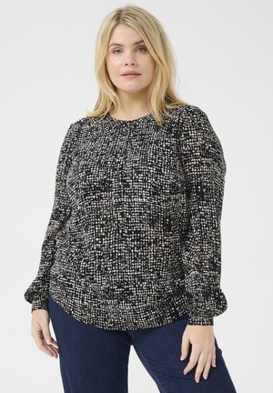 Blouse à manches longues texturée noir et blanc avec un col rond, comportant des motifs irréguliers et des manches froncées, portée sur un jean foncé.