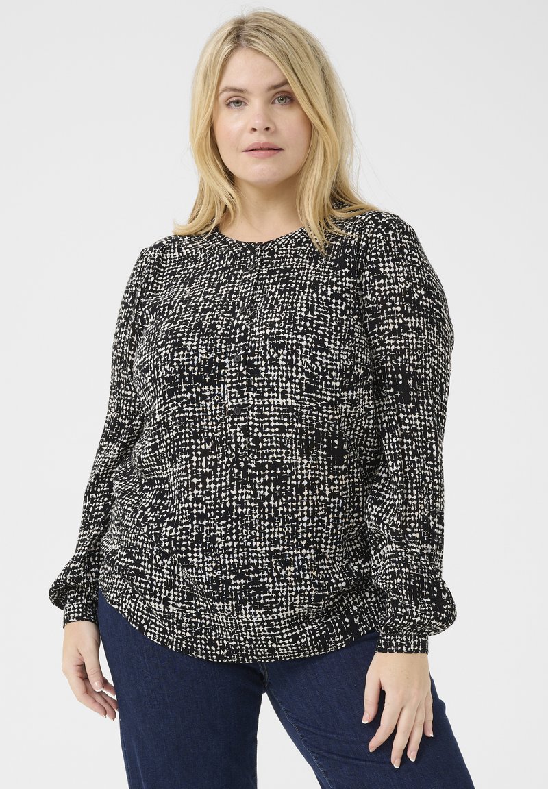 Blusa texturizada en blanco y negro de manga larga con cuello redondo, que presenta patrones irregulares y mangas fruncidas, llevada sobre jeans oscuros.