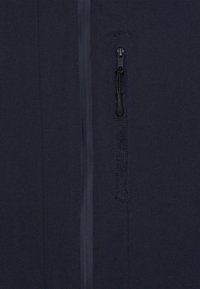 Marinblå mjuk tygjacka med en ficka med dragkedja på vänster sida, med en slät dragkedjestängning och en texturerad detalj nära öppningen.