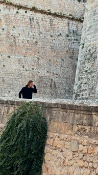 Person in a black sweater stands on a stone wall, surrounded by greenery and a textured stone background with varied colors and patterns.