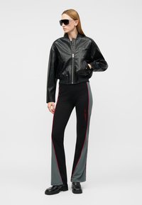 Γυναίκα που φοράει μαύρο υφασμάτινο bomber μπουφάν, μαύρο και γκρι αθλητικό παντελόνι με κόκκινη ρίγα, μαύρα μοκασίνια και μεγάλα μαύρα γυαλιά ηλίου, όρθια.