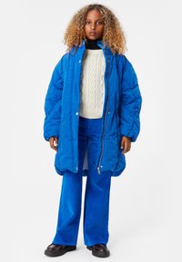 Ljusblå pufferjacka med oversize-passform, tryckknappar och dragkedjestängning, som matchas med blå kordbyxor och en krämfärgad kabelstickad tröja.