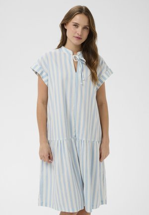 Kvinde iført en løs knælængde kjole med lodrette lyseblå og hvide striber, korte ærmer og bindebånd ved halsudskæringen.