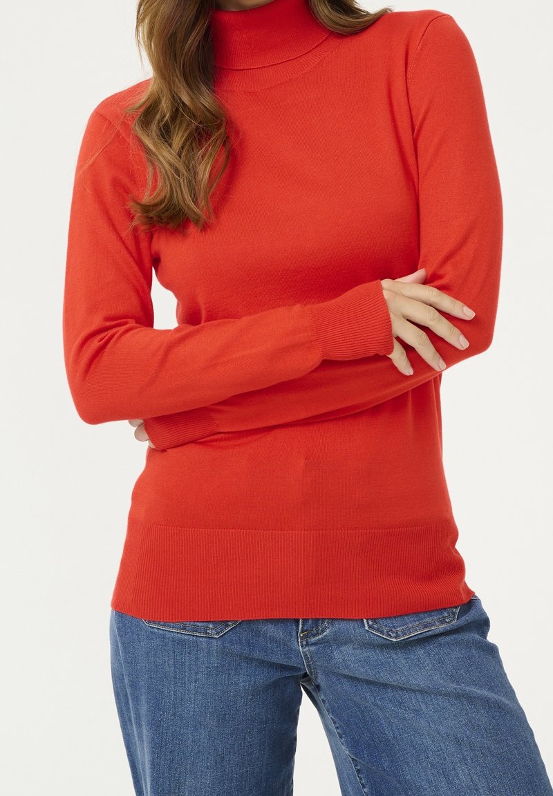 Jersey de cuello alto rojo de manga larga hecho de un material de punto suave, con un diseño ajustado y puños y dobladillo acanalados. Combinado con vaqueros azules.