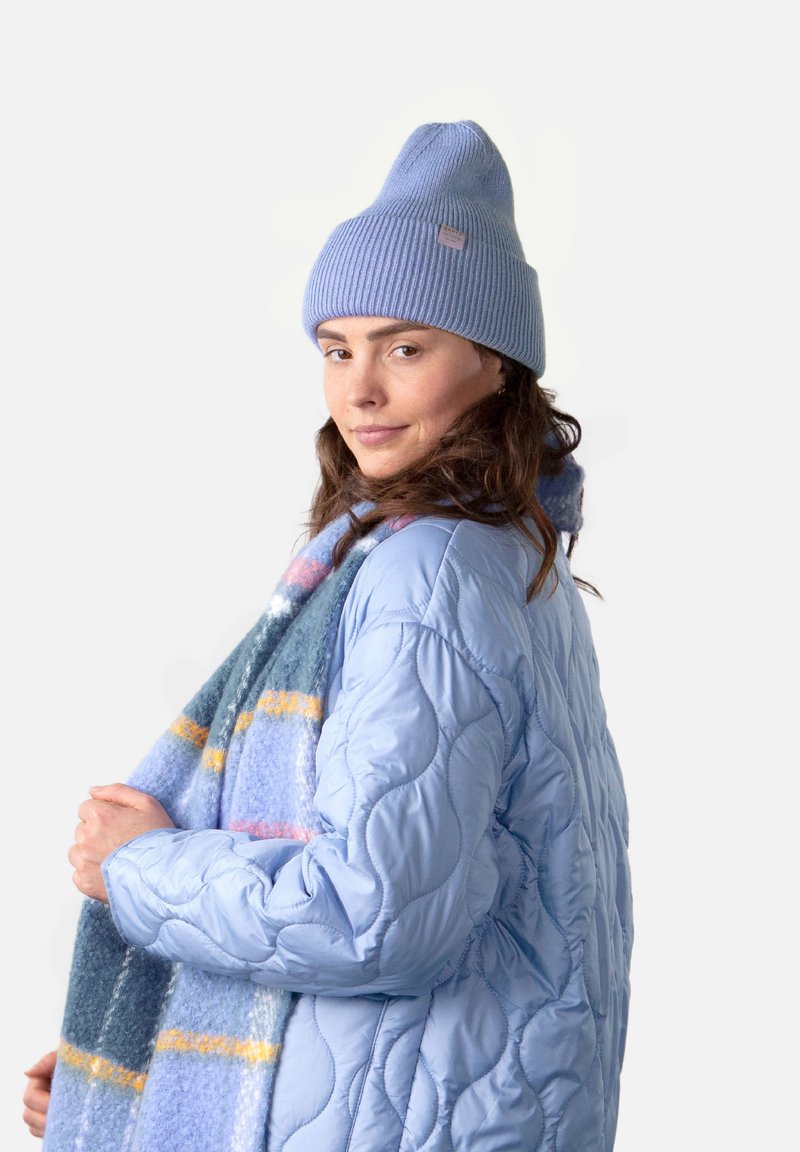 Hellblau gestepptes Puffer-Jackett, geripptes hellblaues Mütze, hält einen mehrfarbigen gestreiften Schal in Blau-, Gelb- und Rosatönen.