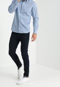  Lichtblauw overhemdd met kleine witte stippen, gecombineerd met donkerblauwe broek en witte sneakers, alles op een effen achtergrond.