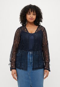 Blusa in pizzo blu navy con maniche lunghe, dotata di chiusura a fiocco al collo e design a strati, indossata sopra un top blu navy e una gonna di jeans.