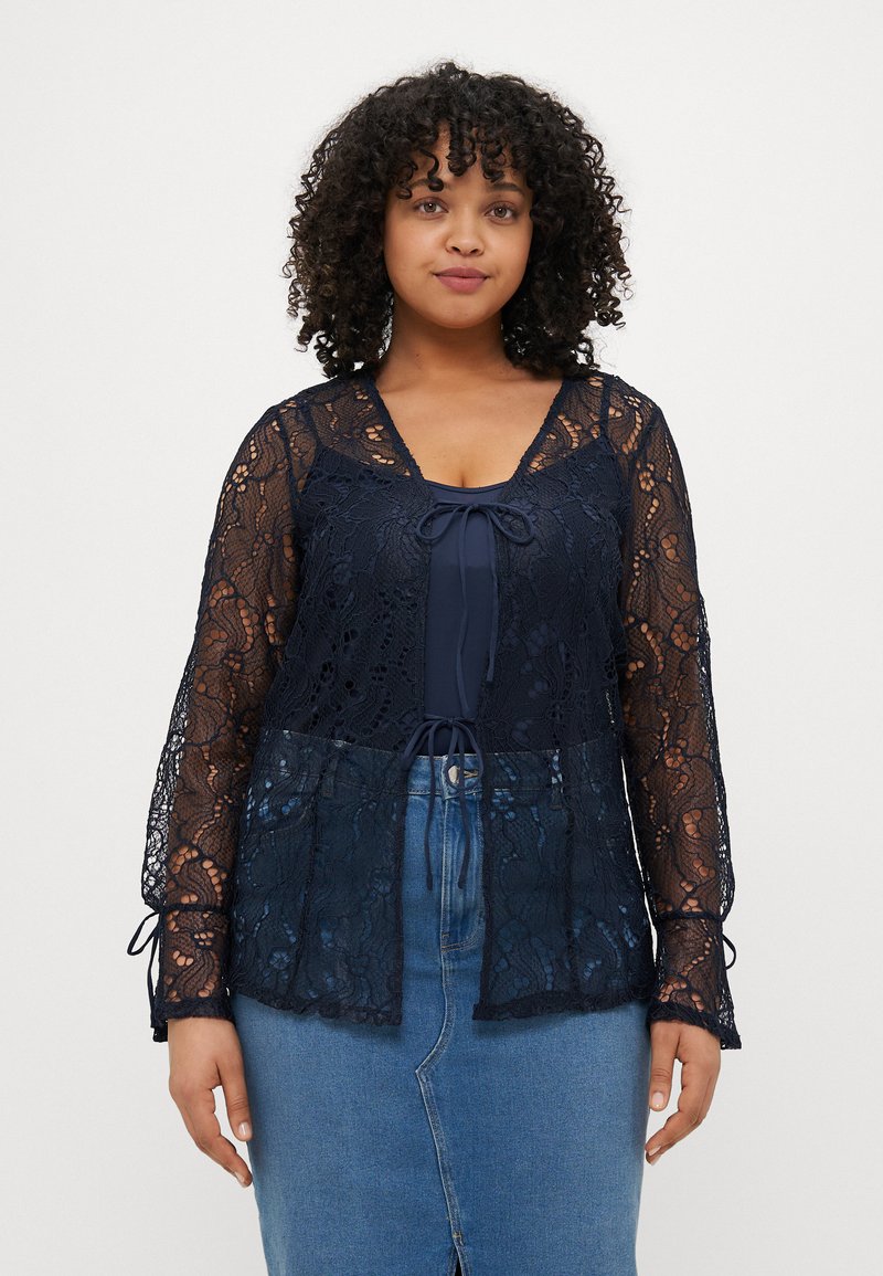 Blusa in pizzo blu navy con maniche lunghe, dotata di chiusura a fiocco al collo e design a strati, indossata sopra un top blu navy e una gonna di jeans.