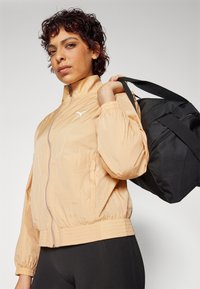 Veste légère de couleur pêche avec un col haut, fermeture éclair, poignets élastiques et ceinture élastique. Sac de sport noir porté sur l'épaule.