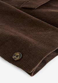 Barna kordbársony anyag, függőleges bordákkal és egy kerek, sötét gombdíszítéssel. Az anyag puha tapintású, hajtogatott szélekkel.