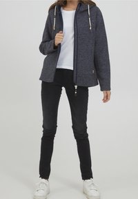 Giacca in pile blu navy con cappuccio, dotata di chiusura a zip, dettagli in perline di legno e tasche frontali, abbinata a pantaloni neri slim-fit e sneakers bianche.
