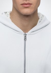 Gros plan d'une personne portant un sweat à capuche blanc zippé, avec une vue partielle des lèvres, du menton et du cou.
