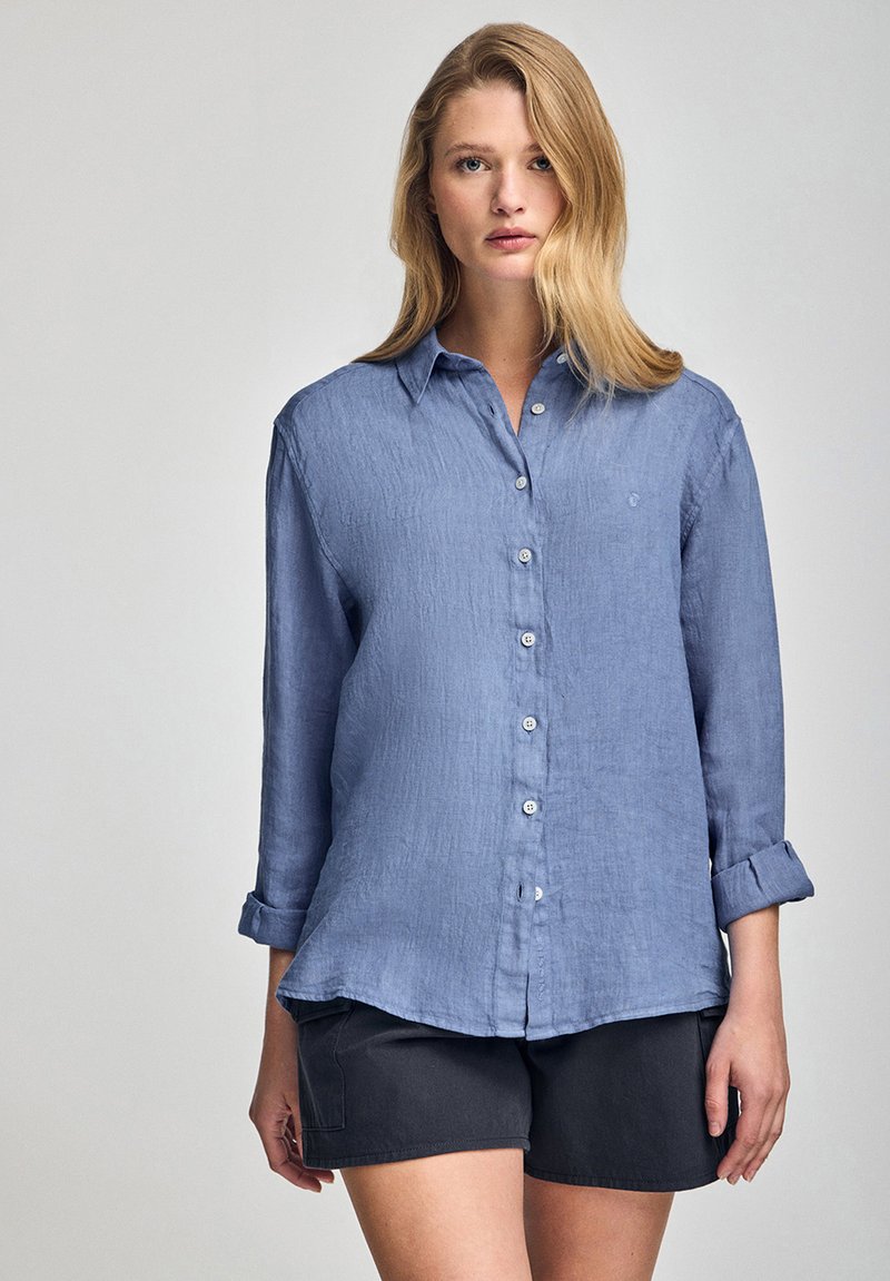 Blå linneskjorta med knapp-ner-stil, lång ärm uppvikt, lätt textur och liten logotyp på vänster bröst. Bärs med mörka shorts.