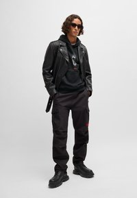 Svart lær bikerjakke over en svart hettegenser med grafisk trykk, cargo-bukser i mørk nyanse, kraftige svarte støvler og solbriller.