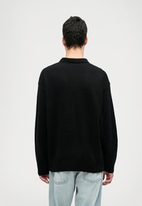 Maglione lavorato a maglia nero con collo a разговину, vestibilità rilassata, polsini e orlo a coste. Indossato con jeans azzurri, visto di spalle.